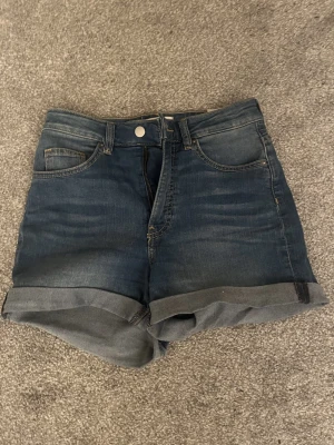 Högmidjade jeansshorts från H&M - Säljer ett par mörkblå högmidjade jeansshorts från H&M i storlek 36. Klassisk femficksmodell med uppvikta benslut och knapp- samt dragkedjestängning. Perfekta till sommaren och lätta att matcha med olika stilar.