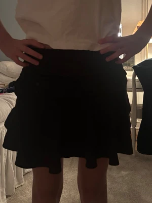 Svart volangkjol med utsvängd form - Säljer en svart kortkjol med volangkant och utsvängd passform. Kjolen har en enkel, stilren look och är perfekt för dig som gillar en flowy och bekväm stil. Passar till många olika outfits och är lätt att matcha.