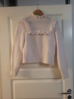 Vit stickad tröja med volang - Supersöt ljusrosa stickad tröja med högt ribbat hals, volangdetaljer över axlarna och ett mönstrat parti framtill. Tröjan har lång ärm och ribbade muddar. Perfekt för dig som vill ha en gullig och trendig look.