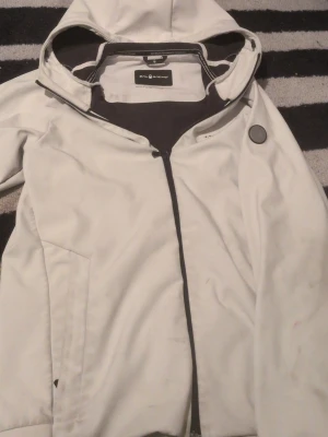 Vit vindjacka från Sail Racing M - Säljer en stilren vit vindjacka från Sail Racing i storlek medium. Jackan har huva, dragkedja framtill och diskret logga på ärmen. Perfekt för blåsiga dagar och enkel att matcha med det mesta. Snygg och sportig look!