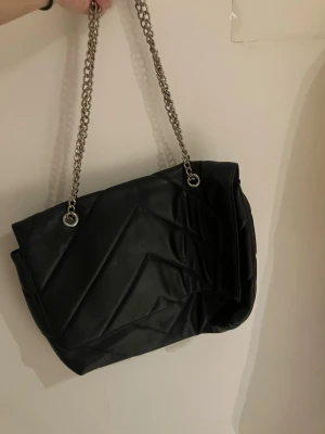 Svart quiltad axelväska från Accessories - Säljer en svart axelväska med quiltad design och snygga kedjehandtag i silver. Väskan har magnetlås och rymligt innerfack. Perfekt för dig som gillar stilrena detaljer och vill ha plats för allt du behöver. Kedjorna ger en cool vibe och väskan är lätt att matcha med olika outfits.