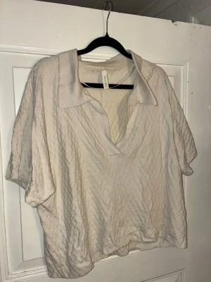 Beige kortärmad topp från Kay/Day - Säljer en beige topp från Kay/Day i storlek S. Toppen har en avslappnad passform, korta ärmar och en bred krage med v-ringad öppning. Materialet är mjukt och har en diskret struktur som ger en cool känsla. Perfekt till jeans eller kjol för en enkel men snygg look.