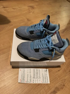 Nike Air Jordan 4 University Blue - Nike Air Jordan 4 University Blue med ovandel i ljusblå mocka, svarta och grå detaljer samt meshpaneler på sidorna. Skorna har klassisk snörning, Jumpman-logga på hälen och en robust sula med svart och grått. Perfekta för dig som vill sticka ut med en ikonisk sneaker. Använda några gånger men är i bra skick