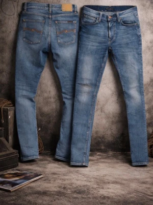 Nudie Jeans Blå  - Säljer ett par snygga jeans från Nudie Jeans i ljusblå tvätt. Jeansen är tillverkade av 100% ekologisk bomull och har klassiska fem fickor, orangea sömmar och Nudie-logga på bakfickan. Perfekta för dig som gillar en smal passform och stilren look. W28 och L32 