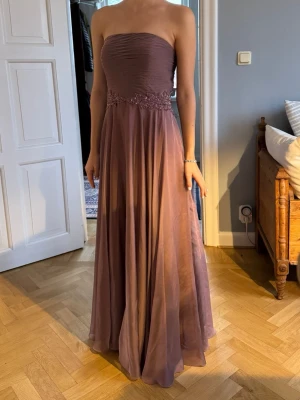 axelbandslös lila balklänning - LILLY - Säljer en elegant axelbandslös långklänning från LILLY i lila med vackra broderade detaljer i midjan och snörning i ryggen. Klänningen har även en chal som kan hängas över axlarna eller runt halsen. Klänningen har ett svepande fall och är tillverkad i ett lätt, skirt material som ger en lyxig känsla. Stirlek 40 men då den är en korset så passar den många fler. (Ca 46-36) Aldrig använd, Danish design, köpt i Köpenhamn, nypris 3900kr