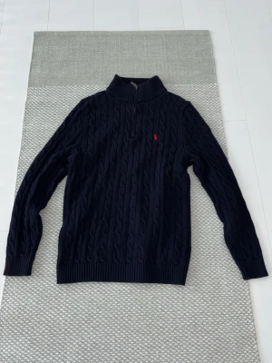 Marinblå stickad polotröja Polo Ralph Lauren - Säljer en marinblå stickad polotröja från Polo Ralph Lauren i storlek M. Tröjan har kabelstickat mönster, ribbad krage och half zip-dragkedja. Klassisk röd broderad logga på bröstet. Perfekt för dig som gillar stilrena och tidlösa plagg.