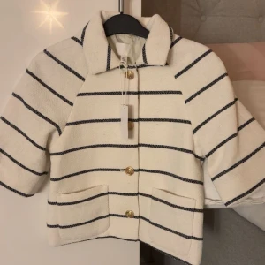 Randig vit kavajjacka från H&M - Snygg vit kavajjacka med svarta horisontella ränder och guldiga knappar framtill. Jackan har krage, två fickor och är tillverkad i ett strukturerat tyg som ger en cool vibe. Perfekt för dig som gillar klassisk stil med en twist.