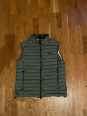 Grön dunväst från Colmar - Säljer en snygg grön dunväst från Colmar med quiltad design och hög krage. Västen har två dragkedjeförsedda fickor framtill och en diskret logga på bröstet. Insidan är ljusgrå och västen stängs med dragkedja. Perfekt för lager-på-lager under kyliga dagar.