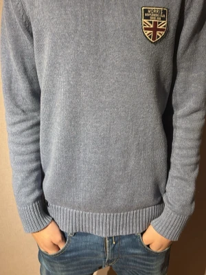 Ljusblå stickad tröja från Morris - Säljer en ljusblå stickad tröja från Morris Gents i storlek Large. Tröjan har rund hals, ribbade muddar och en snygg patch med brittisk flagga på bröstet. Perfekt till jeans för en clean och stilren look.