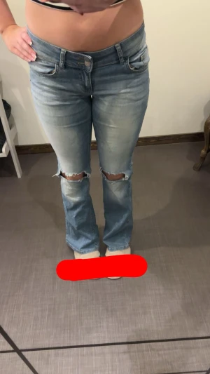 Ljusblå bootcut jeans med slitningar - Säljer ett par ljusblå bootcut jeans med slitna hål över knäna och snygga tvättade detaljer. Jeansen har låg midja, klassiska fem fickor och bakfickor med lock och knapp. Perfekta för dig som gillar en avslappnad och trendig look.