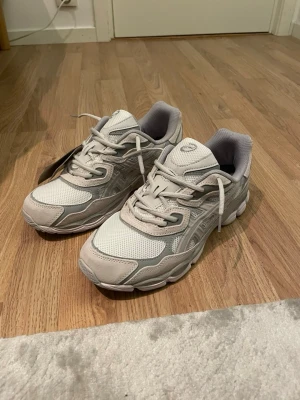 Vita och grå ASICS sneakers - Säljer ett par fräscha ASICS sneakers i vitt och ljusgrått med meshpaneler och detaljer i syntet och mocka. Skorna har snörning, Ortholite innersula och en chunky sula som ger skön dämpning. Perfekta för dig som gillar sportig och clean stil.