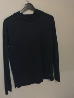 Svart golf hoddie med Luca tunn tröja.  - En enkel svart hoodie med huva och långa ärmar. Tröjan har en clean look utan synliga tryck eller detaljer och är perfekt för dig som gillar minimalistisk stil. Passar till både chill och streetwear.