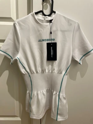 Vit Kim topp från J.Lindeberg - HELT NY Snygg vit topp från J.Lindeberg med gröna detaljer och ribbad midja. Modellen har korta ärmar, rund hals och dragkedja i ryggen. Perfekt för en sportig och fräsch look, tillverkad i ett mjukt och stretchigt material.