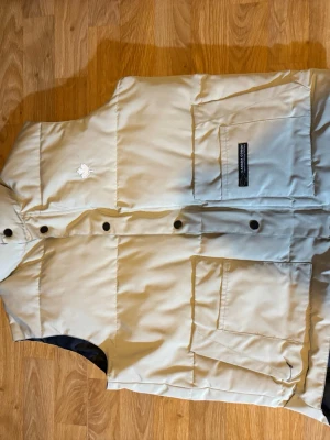 Beige dunväst från Canada Goose - Säljer en beige dunväst från Canada Goose med stora fickor framtill och tryckknappar. Västen har en diskret logga med lönnlöv på bröstet och svart innerfoder. Perfekt för kalla dagar när du vill hålla stilen.