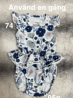 Blommig body från Polarn O. Pyret - Supersöt blommig body i vitt och blått från Polarn O. Pyret. Volangdetaljer vid axlar och midja, tryckknappar på axeln och i grenen för enkel påklädning. Perfekt för vår och sommar!🌸Använd en gång. Storlek 74 