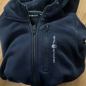 Svart Sail Racing jacka med huva - Svart jacka från Sail Racing med huva och dragkedja framtill. Jackan har en bröstficka med dragkedja och broderad logga på bröstet. Mjuk insida och sportig look, perfekt för dig som gillar stilrena och funktionella plagg.