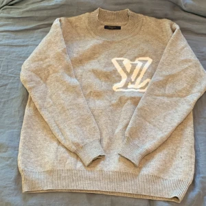 Grå stickad tröja Louis Vuitton - Snygg grå stickad tröja från Louis Vuitton med stort vitt LV-logo på både fram- och baksidan. Tröjan har rund halsringning, ribbade muddar och är långärmad. Perfekt för dig som vill ha en exklusiv och stilren look.