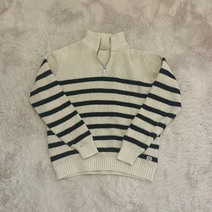 Randig stickad half zip-tröja - Snygg stickad tröja i beige med svarta ränder och half zip-dragkedja vid halsen. Tröjan har ribbade muddar och hög krage, perfekt för lager-på-lager. Klassisk och chill stil som funkar året runt.