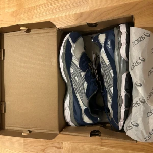 Blå och silver Asics sneakers - Snygga Asics sneakers i marinblått och silver med vita och grå detaljer. Skorna har en sportig design med mesh och syntetmaterial, samt snörning och platt sula. Perfekta för dig som gillar stilrena och bekväma sneakers med coola kontraster.