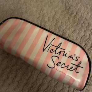 Randig necessär från Victoria's Secret - Snygg necessär från Victoria's Secret med rosa och vita ränder på utsidan och prickigt svart foder. Väskan har en praktisk dragkedja och är perfekt för smink eller småprylar. Den är tillverkad i syntetmaterial och har en stilren, mjuk form.