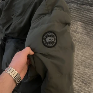 Grå dunjacka från Canada Goose - Säljer en grå dunjacka från Canada Goose med klassisk patch på ärmen. Jackan har huva, stora fickor och stängs med både dragkedja och knappar. Perfekt för kalla dagar och riktigt snygg streetstil.Jag har använt den bara ett par gånger men jag säljer den för jag ska köpa ny jacka. Den är storlek L men det passar som storlek M också.