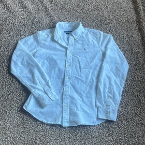 Vit slim skjorta från Ralph Lauren - Vit skjorta från Ralph Lauren med klassisk krage och broderad logga på bröstet. Super slim fit för en tajt och modern siluett. Långärmad med knappar framtill och vid ärmslut. Tillverkad i mjuk bomull som känns skön mot huden.  Passar dig mellan 160-170. !!!Litet hål i armen men syns inte vid användning!!! Har du fler frågor? Tveka inte skriv!! MVH SWISSWEAR 🇨🇭🍾🏔️