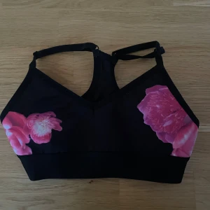 Svart blommig linnetopp Gina Tricot XS - Svart linnetopp från Gina Tricot med tunna axelband och stora rosa blommor på framsidan. Toppen har en sportig vibe och bred resår under bysten för extra stöd. Perfekt för dig som gillar färgstarka detaljer och vill sticka ut.