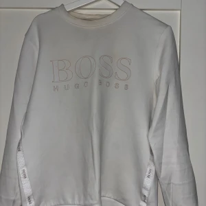Vit sweatshirt från Hugo Boss - Säljer en stilren vit sweatshirt från Hugo Boss med logga framtill och dekorativa band med BOSS-tryck längs sidorna. Tröjan har rund halsringning och långa ärmar, perfekt för en clean och trendig look.