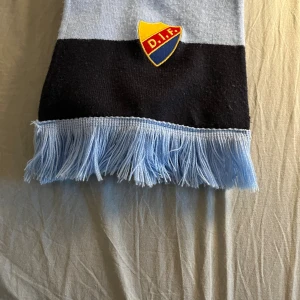 Randig DIF halsduk med fransar - Snygg halsduk från Djurgårdens IF med breda marinblå och ljusblå ränder, broderat DIF-märke och fransar i ljusblått i ändarna. Perfekt för att visa ditt stöd på matchen eller bara hålla dig varm. Lång och mjuk, passar dig som vill sticka ut på läktaren.