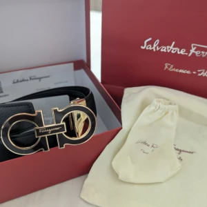 Ferragamo Bälte - Snyggt svart bälte från Salvatore Ferragamo med exklusivt Gancini-spänne i guld och svart. Bältet är tillverkat i äkta skinn och har en stilren, klassisk form som lyfter din outfit. Kommer med originalförpackning och dustbag.