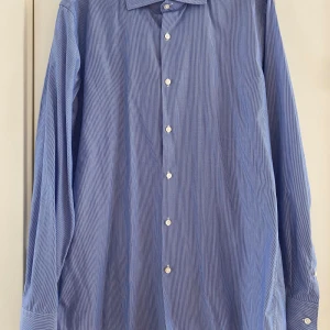 Blå randig skjorta från Hugo Boss - Snygg blå skjorta från Hugo Boss i slim fit-modell. Skjortan har tunna vita ränder, klassisk krage och vita knappar. Tillverkad i bomull och har långa ärmar med manschett. Perfekt för dig som gillar stilrena och fräscha plagg. Mått Över bröstet: ca 46 cm Längd från axeln och ner: ca 76 cm  Material: 100% bomull  Kommer från ett djur- och rökfritt hem.