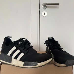 Svarta Adidas NMD sneakers strl 40 2/3 - Adidas NMD sneakers i svart med vita ränder på sidorna och en vit, texturerad Boost-sula. Skorna har snörning, platt sula och detaljer i syntetmaterial. Ikonisk NMD-design med svart och vit färgkombination, perfekt för dig som gillar streetwear och sportig stil.