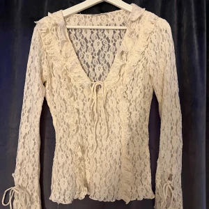 Spetsblus med volang från Zara - Romantisk spetsblus från Zara i ljusbeige med v-ringning, volangkant och knyt framtill. Blusen har långa ärmar med knyt vid ärmslut och är helt genombruten i spets. Perfekt för dig som gillar vintagevibbar och detaljer.💓
