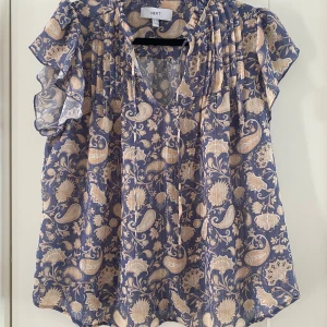 Blommig blus från Next i blått och beige - Säljer en luftig blus från Next med blommigt och paisleymönster i beige på blå botten. Blusen har v-ringning, volangiga korta ärmar och är tillverkad i ett tunt, lätt material. Perfekt för dig som gillar boho-vibes och snygga detaljer. Mått Över bröstet: ca 55 cm Längd från axeln och ner: ca 64  100% polyester  Kommer från ett djur och rökfritt hem.