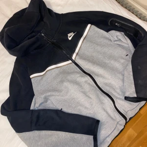 Nike hoodie i svart och grå med dragkedja - Nike hoodie med huva i svart och ljusgrått, med vit logga på bröstet och snygga kontrastlinjer. 