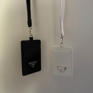 Pr4da svarta och vita korthållare - Två stilrena korthållare från Pr4da, en i svart och en i vitt skinn. Båda har avtagbara handledsremmar och Pr4das ikoniska metallogo framtill. Perfekt för kort och ID med transparent fönster på baksidan. Snygg accessoar för dig som gillar lyxiga detaljer.