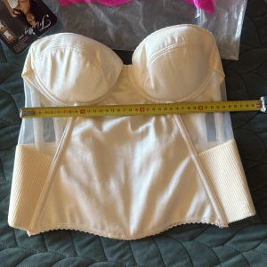 Champagnefärgad korsett från FLattering me - Snygg champagnefärgad korsett från Flattering me med vadderade kupor och transparenta meshpaneler på sidorna. Toppen har breda ribbade partier, hakknäppning bak och en glansig satinliknande finish. Perfekt för att skapa en cool och elegant look.