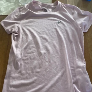 Ljusrosa Asics t-shirt - Säljer en ljusrosa t-shirt från Asics med korta ärmar och rund hals. T-shirten har en diskret Asics-logga på bröstet och är tillverkad i mjukt, lätt polyester som andas. Perfekt för träning eller chill dagar.