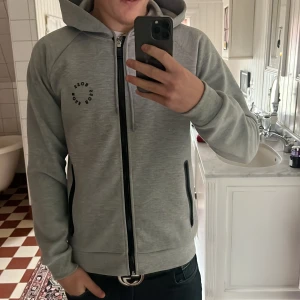Grå hoodie från BOSS med dragkedja - Snygg ljusgrå hoodie från BOSS med svart dragkedja och svarta detaljer vid fickorna. Tröjan har huva med snörning och tryckt BOSS-logga på bröstet. Perfekt för chill dagar och har en modern, clean look. Materialet är mjukt och känns skönt mot huden. Tröjan är använd max 2 gånger och är i väldigt fint skick! 