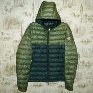 Grön Moncler dunjacka med huva - Snygg dunjacka från Moncler i två nyanser av grönt. Jackan har quiltad design, huva och dragkedja framtill. Klassisk Moncler-logga på ärmen. Perfekt för dig som vill ha en lätt och stilren jacka med streetkänsla.