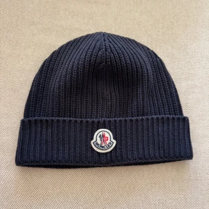 Mörkblå Moncler mössa med logga - Säljer en mörkblå ribbstickad Moncler mössa i ull med uppvikt kant. Framtill sitter Monclers klassiska broderade logotyp i vitt, rött och blått samt en stor vit broderad MC-logga på andra sidan. Grym accessoar för kalla dagar.