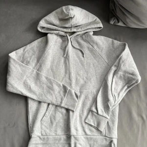 Ljusgrå hoodie med huva och ficka - Säljer en stilren ljusgrå hoodie med huva och dragsnören. Tröjan har en klassisk känguruficka framtill och långa ärmar med breda muddar. Perfekt för dig som gillar en enkel och avslappnad look.