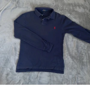 Marinblå långärmad pikétröja Polo Ralph Lauren - Snygg marinblå långärmad pikétröja från Polo Ralph Lauren med klassisk krage och två knappar. Tröjan har det ikoniska röda Polo-logot broderat på bröstet och är gjord i mjuk bomull. Perfekt för dig som gillar stilrena och tidlösa plagg.