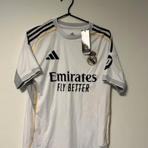 Real Madrid vit matchtröja  - Säljer vit Real Madrid fotbollströja från Adidas med svarta ränder på axlarna och gula detaljer. Broderat klubbmärke på bröstet och tryckt sponsortext. Tillverkad i lätt polyester, perfekt för träning eller match.