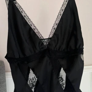 Svart satin topp med spets - Säljer en svart topp i satin med djup v-ringning och dekorativa spetsdetaljer längs urringningen och midjan. Topp har tunna axelband och transparenta spetsinslag med blommigt mönster som ger en extra lyxig känsla.