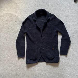 Mörkblå stickad kofta från LCW Casual - Säljer en mörkblå stickad kofta från LCW Casual med snygg krage och två stora framfickor. Koftan har ett diskret mönster, knappar framtill och en chill, klassisk look. Perfekt för lager-på-lager och att hålla sig varm under kyliga dagar.