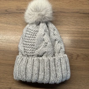 Grå stickad mössa med fluffig boll H&M - Snygg grå stickad mössa från H&M med bred uppvikt kant och en stor fluffig boll på toppen. Flätstickad design som ger extra stilpoäng och mysig känsla. Perfekt för kalla dagar och enkel att matcha med din vinterjacka.