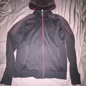 Svart hoodie från Hugo Boss med röd dragkedja - Svart hoodie från Hugo Boss med röda detaljer och dragkedja framtill. Tröjan har huva med snörning, två fickor och diskret Boss-logga på bröstet. Perfekt för en chill och stilren look.