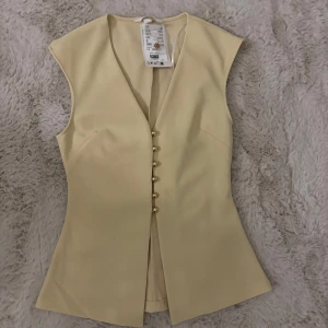 Beige/ljusgul väst med guldknappar - Stilren beige väst från H&M med figurnära passform och snygga guldknappar framtill. Västen har v-ringning och är ärmlös, perfekt för att lyfta din outfit med en clean look.