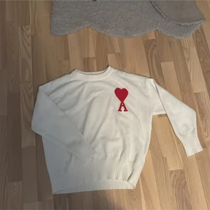 Vit stickad tröja från Ami Paris - Säljer en snygg stickad tröja från Ami Paris med deras ikoniska röda hjärta och A på bröstet. Tröjan har rund halsringning, långa ärmar och ribbade muddar. Perfekt för dig som vill ha en stilren men ändå unik look.STORLEK M PASSAR DEM FRÅN 175-180